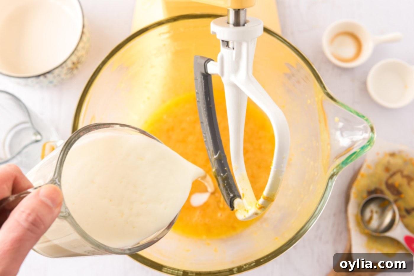 Pouring buttermilk into the orange-infused egg mixture in a stand mixer, demonstrating a key step in cupcake batter preparation.