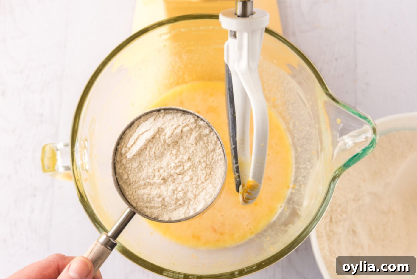 Adding a cup of flour mixture to a bowl of orange cupcake batter in a stand mixer, illustrating the final combining step for the batter.