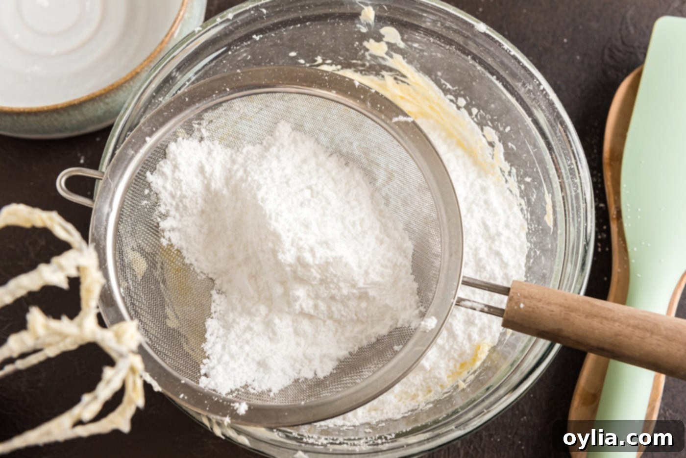 Sifted powdered sugar is being carefully added to the beaten butter mixture in a bowl, ready for further mixing to create the frosting.