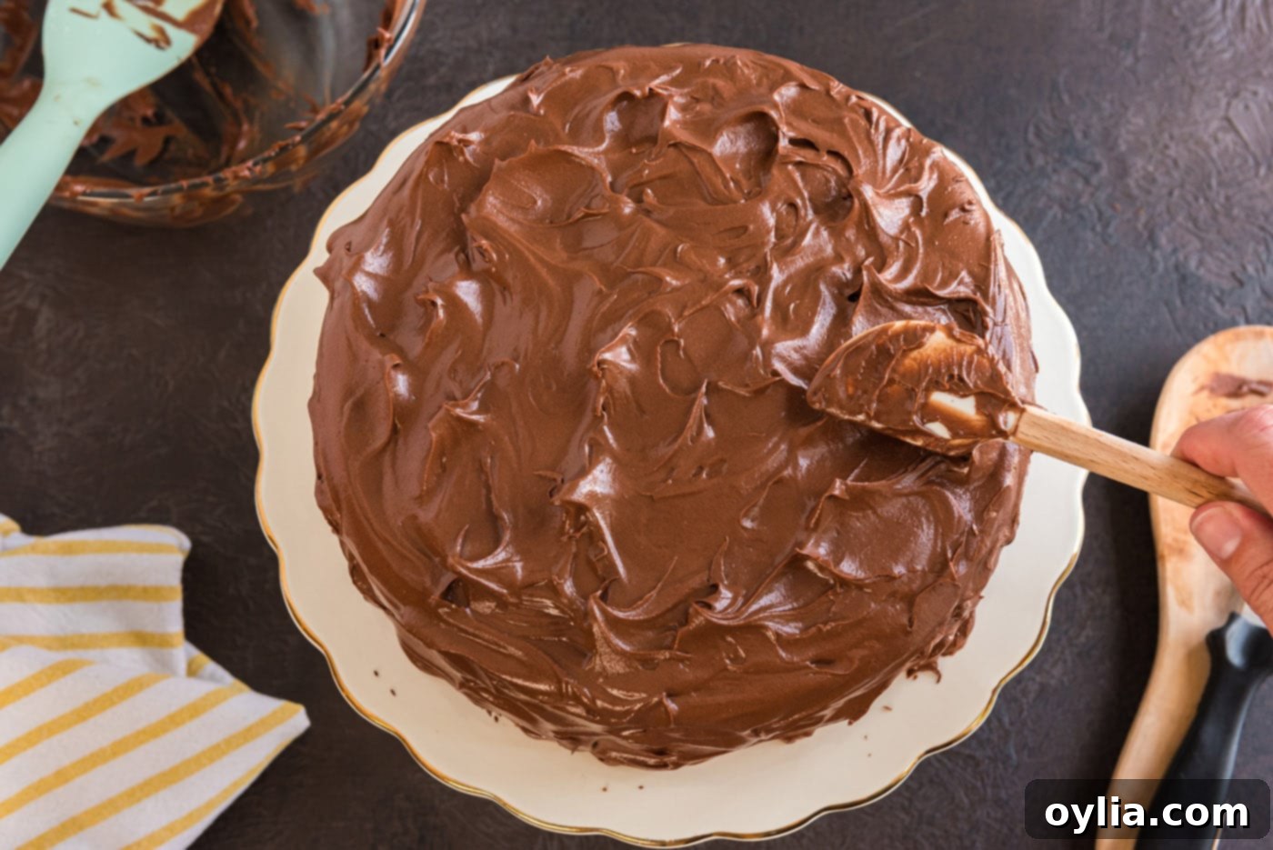 A fully frosted Nigella's Chocolate Fudge Cake sits on a cake stand, showcasing its smooth, rich chocolate fudge icing covering both layers and sides.