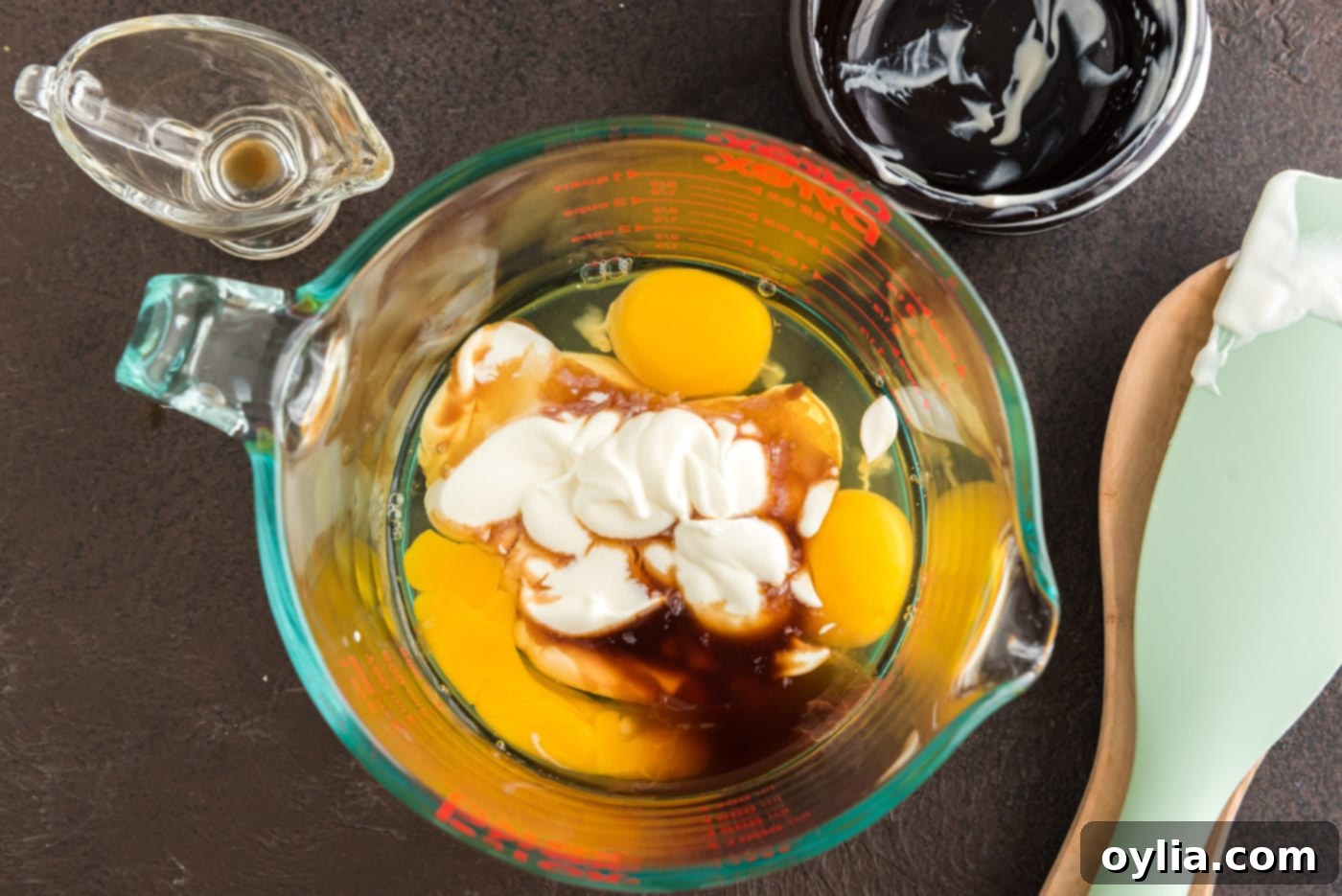 Three large eggs, a dollop of sour cream, and a splash of vanilla extract are neatly placed in a glass bowl, ready to be whisked.