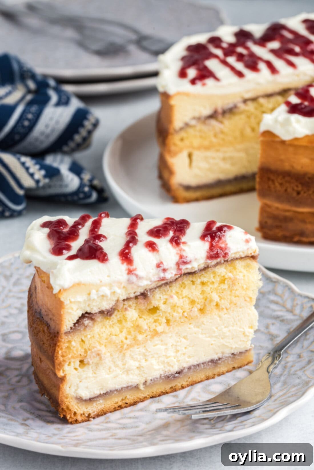 A beautiful slice of English Trifle Cheesecake showing distinct layers of cake, raspberry, and cheesecake, topped with whipped cream and raspberry drizzle.