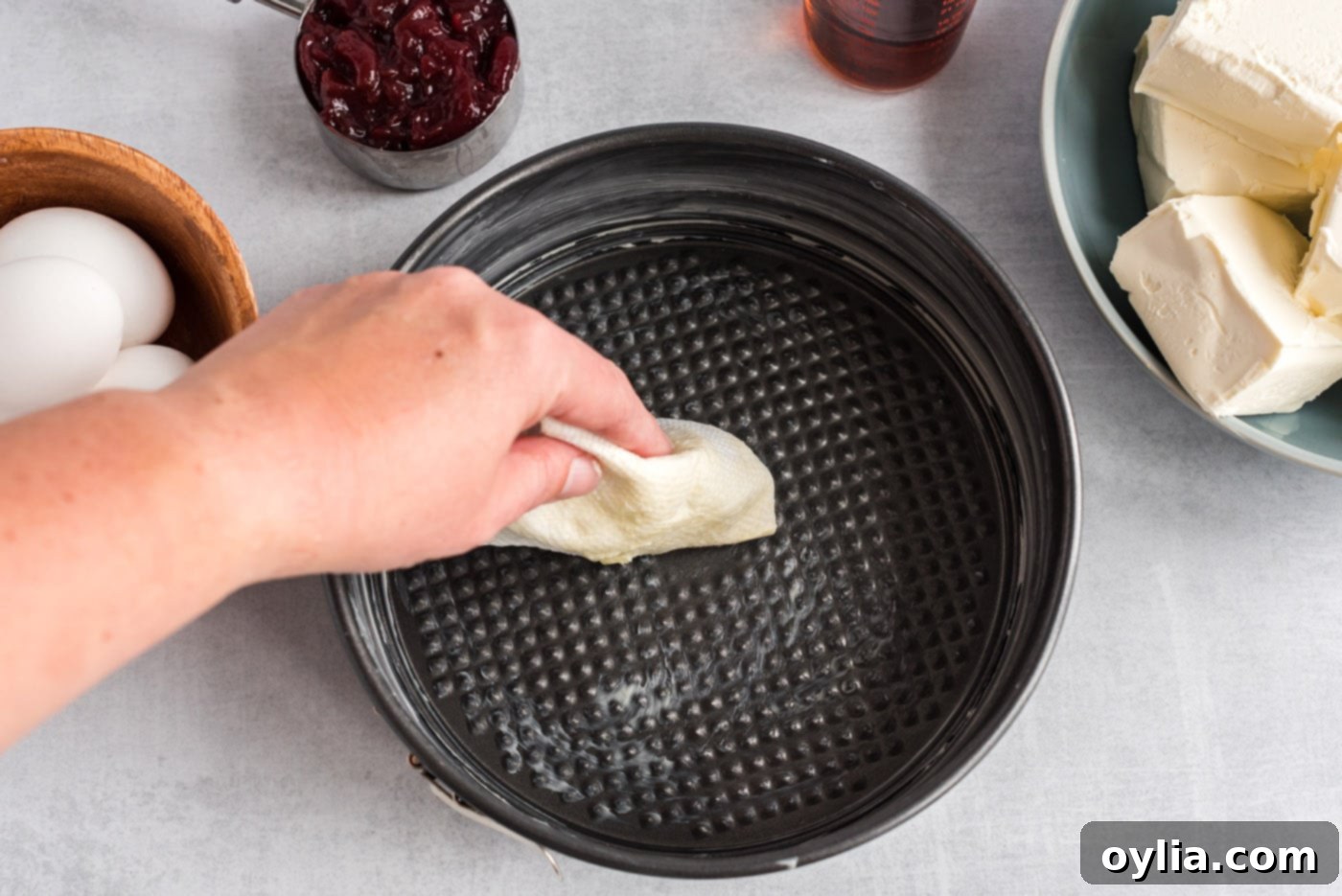A hand buttering the inside of a 9-inch springform pan, preparing it for the cheesecake layers.