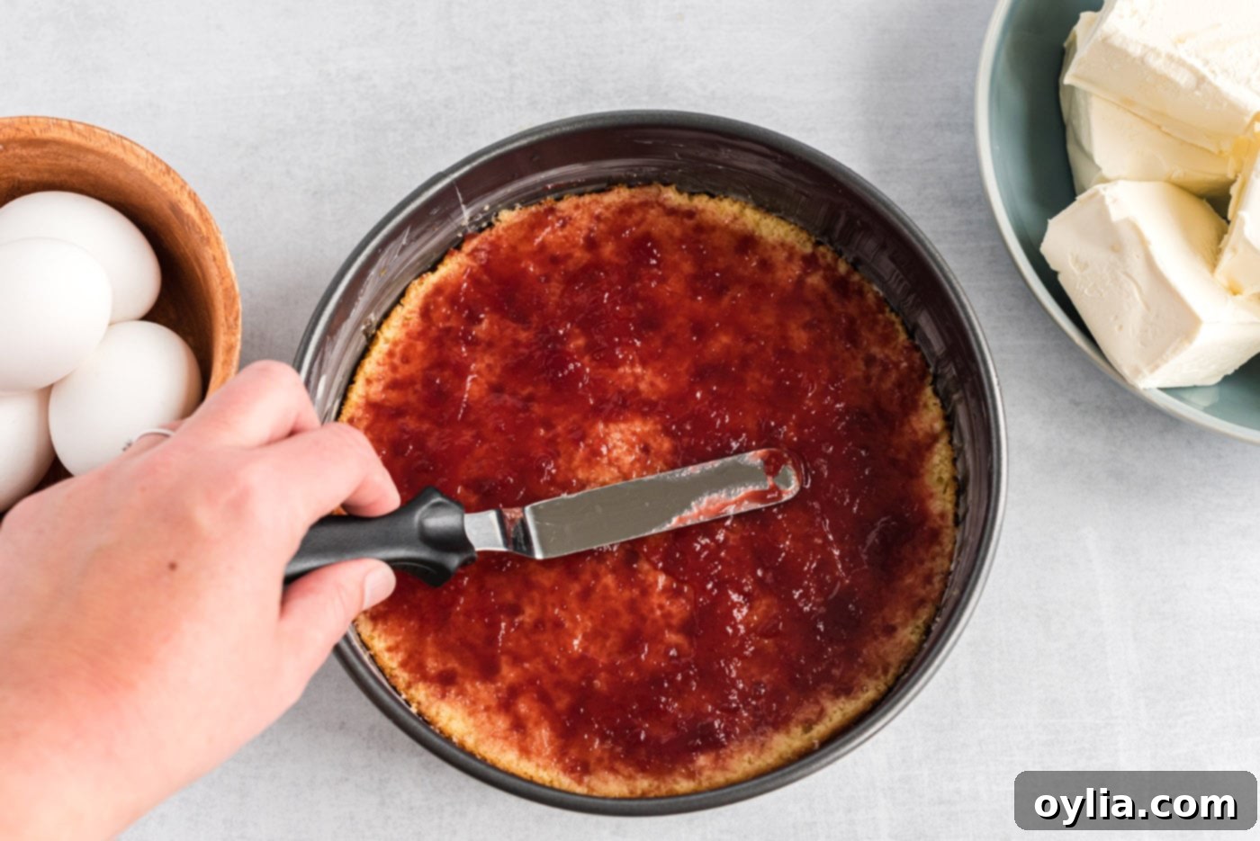 An icing spatula spreading a generous layer of red raspberry preserves over a yellow cake layer.