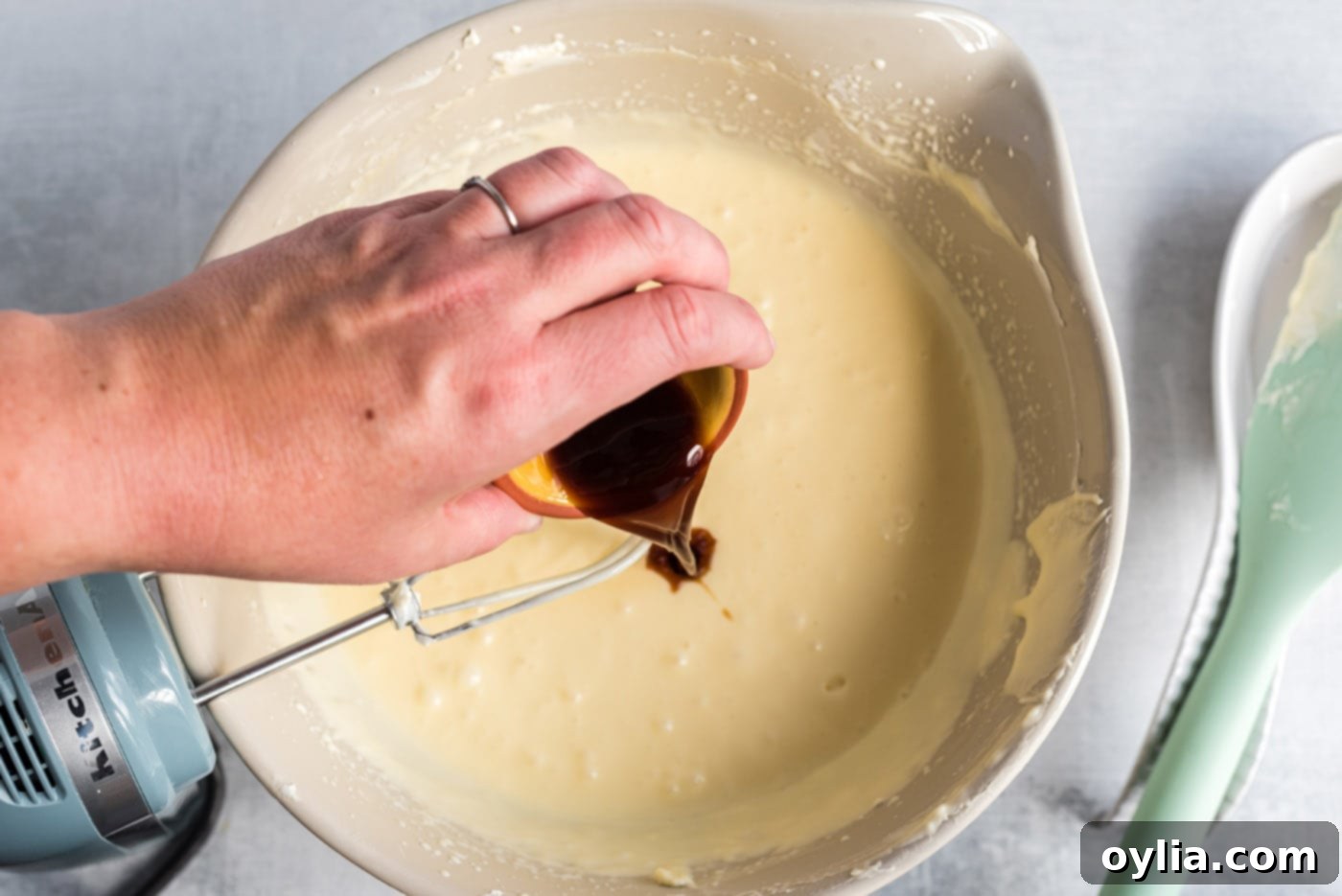 A hand pouring vanilla extract into a bowl of creamy cheesecake mixture being blended by a hand mixer.