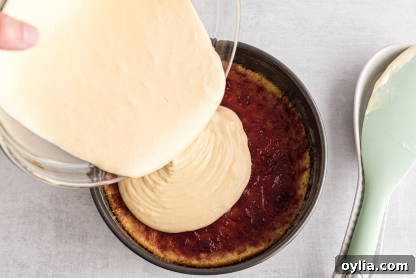 A rich cheesecake mixture being poured over a layer of raspberry preserves and yellow cake in a springform pan.