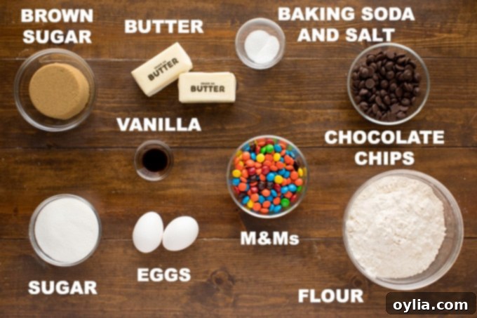 A collection of ingredients laid out for baking chocolate chip M&M cookies, including flour, sugars, eggs, butter, chocolate chips, and M&Ms.