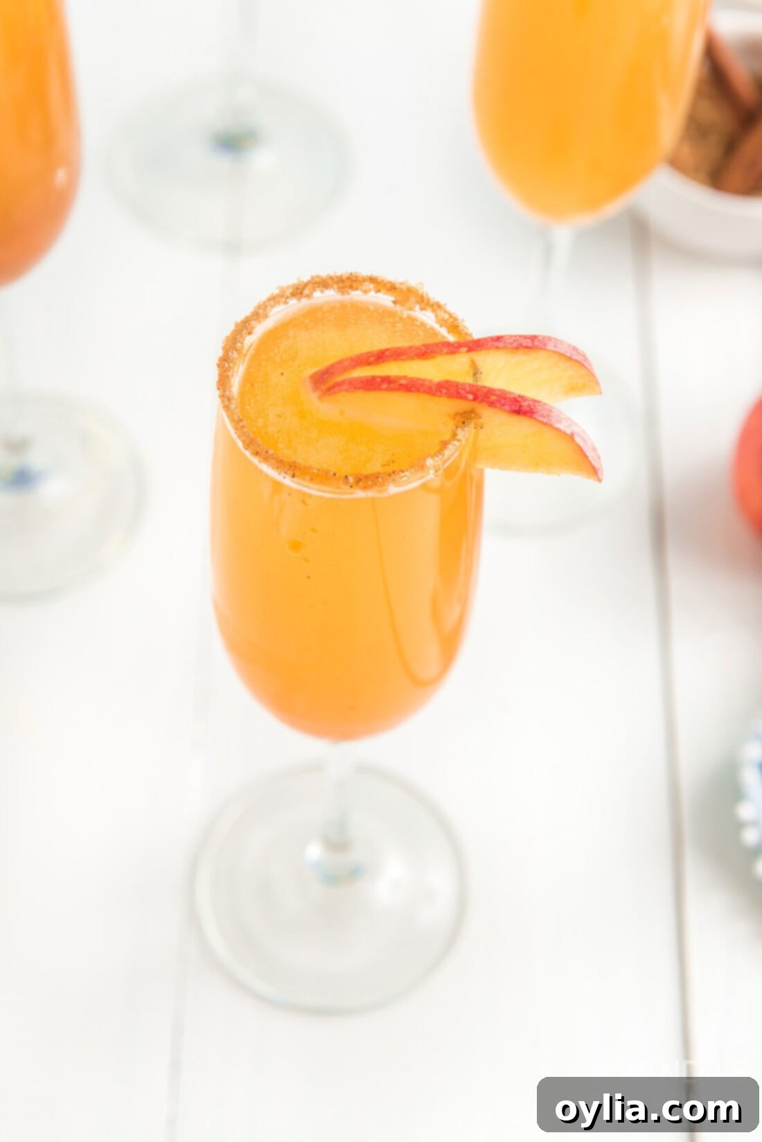 A single glass of caramel apple mimosa, garnished with an apple slice, standing on a white surface.