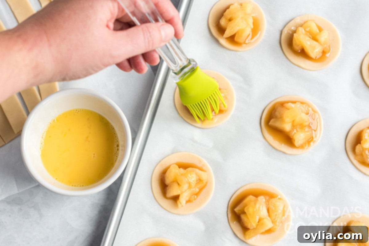 A pastry brush applying egg wash to the edges of a pie dough circle on a baking sheet, preparing it for the lattice top.