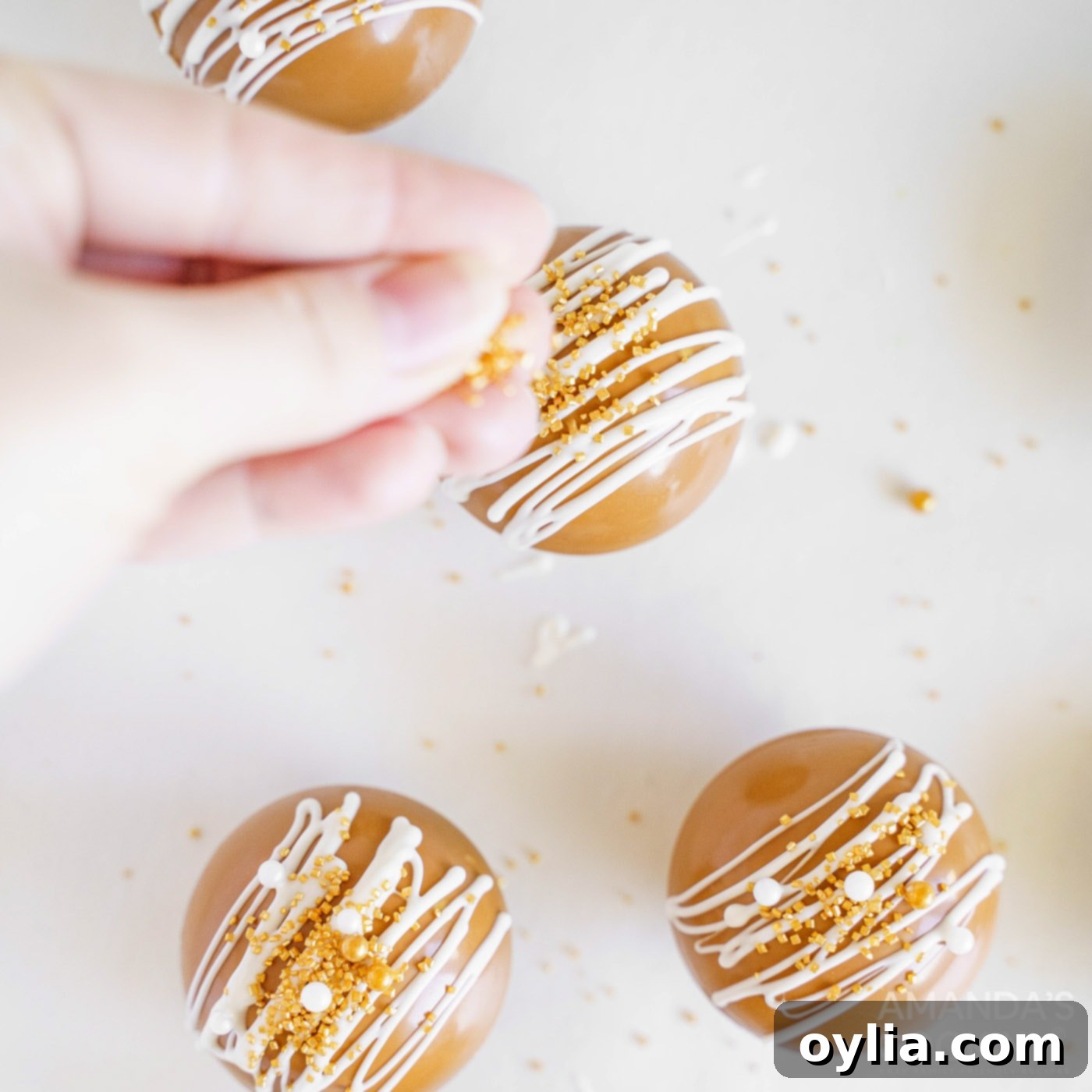 Sprinkles being added to a caramel hot cocoa bomb with white chocolate drizzle, completing the decoration.