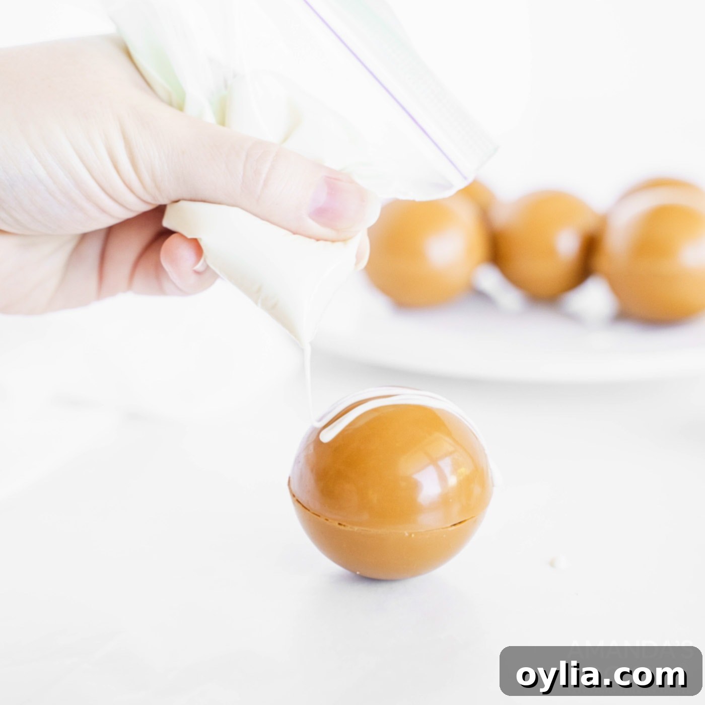 A hand expertly drizzling melted white chocolate over a caramel hot cocoa bomb, creating a decorative pattern.