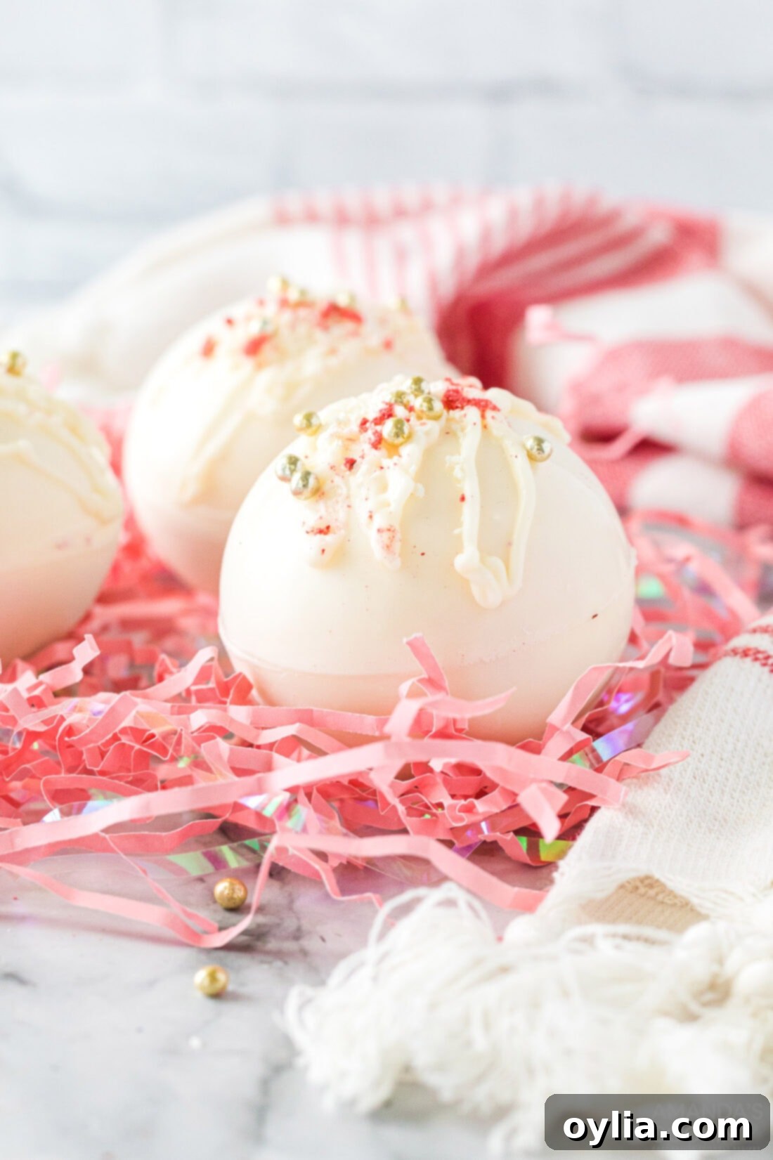 Indulgent Strawberry White Chocolate Hot Cocoa Bombs 2 Beautiful white chocolate hot cocoa bombs with strawberry powder and sprinkles, resting on pink shredded paper, ready for gifting or enjoying.