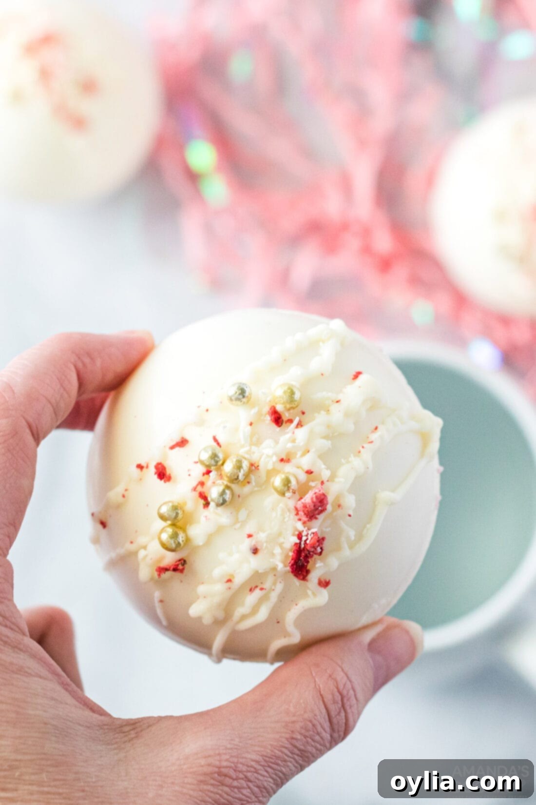 Indulgent Strawberry White Chocolate Hot Cocoa Bombs 3 A hand holding a homemade strawberry white chocolate hot cocoa bomb over a ceramic mug, ready to be dropped in hot milk.