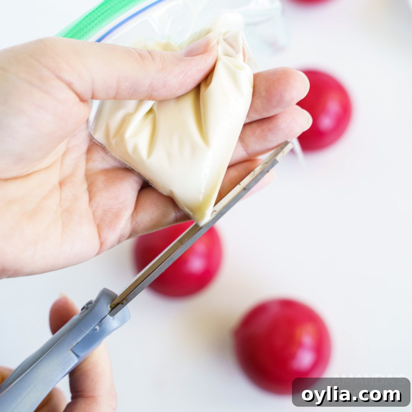 A hand is carefully snipping a tiny corner off a zip-top plastic bag filled with melted white candy, preparing for drizzling.