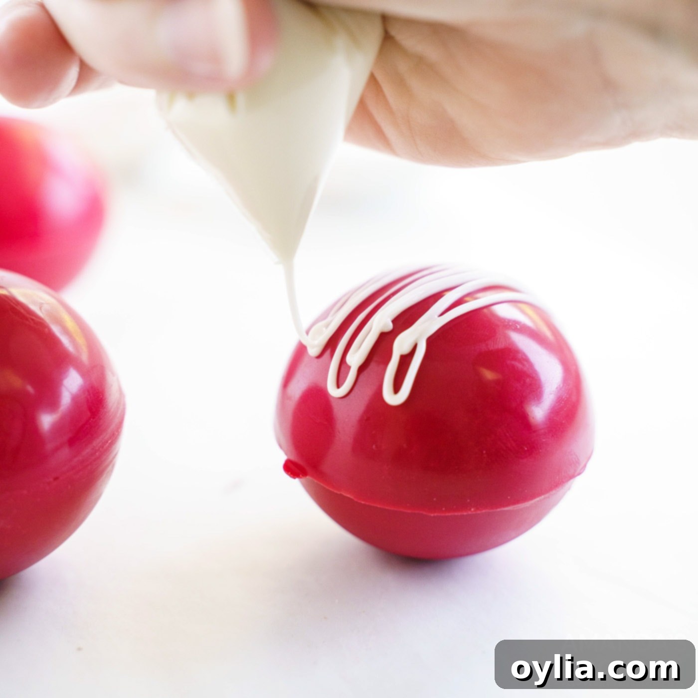 Melted white candy is being drizzled decoratively over the top of a red velvet hot cocoa bomb.
