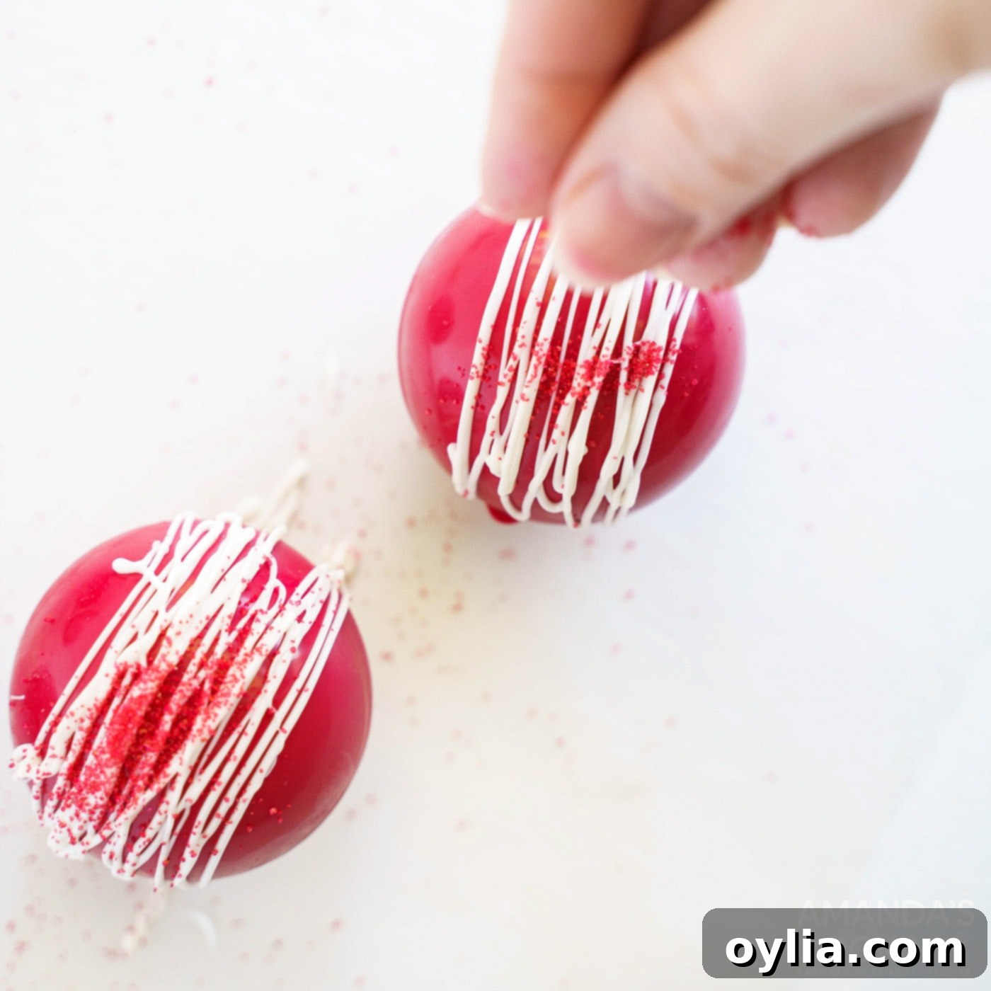 Additional sprinkles are being added to a decorated hot cocoa bomb while the white candy drizzle is still wet.
