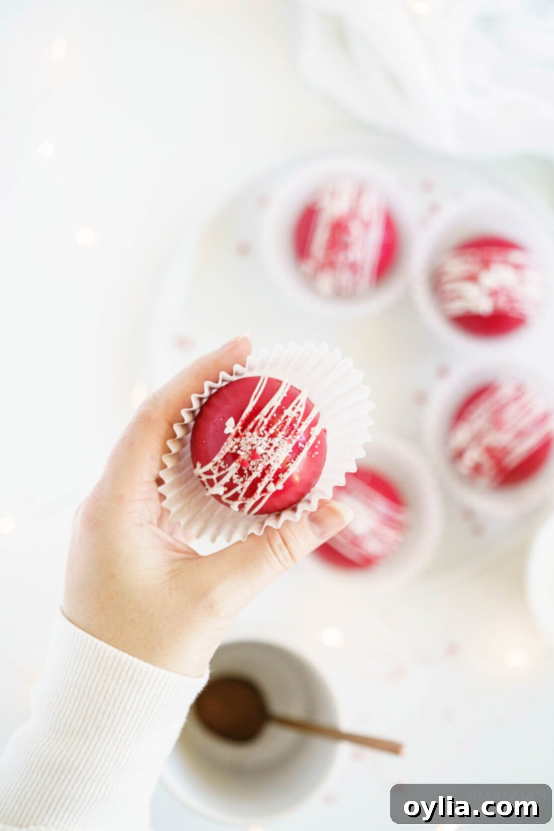 Woman holding a single Red Velvet Hot Cocoa Bomb in a paper liner, showcasing its perfect spherical shape and decorative drizzle.
