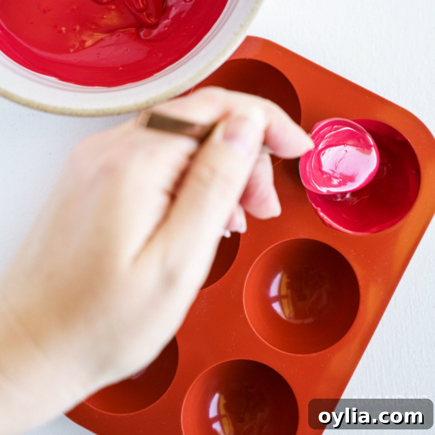 Melted red candy is being carefully spooned into individual silicone sphere molds, forming the outer shells of the hot cocoa bombs.