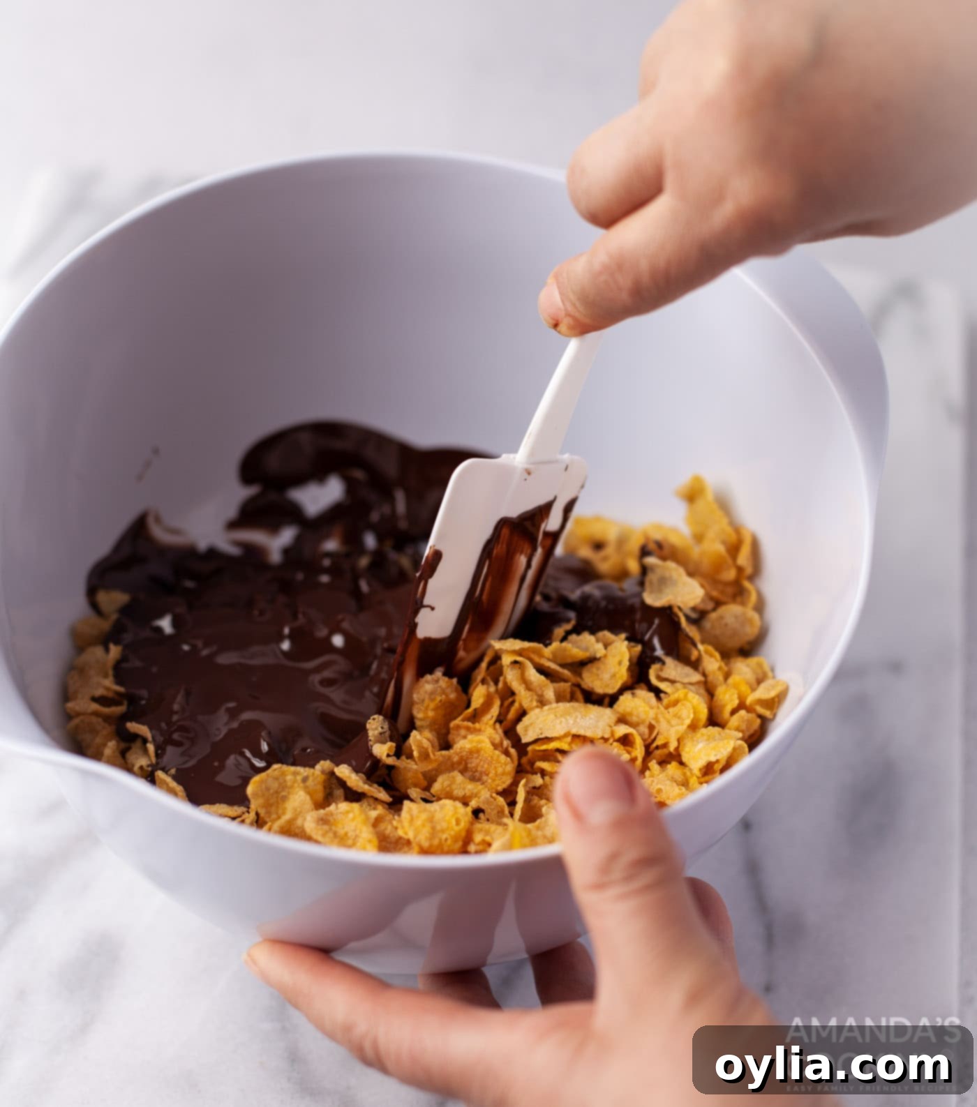 A bowl of cornflakes being carefully mixed with dark melted chocolate using a rubber spatula.