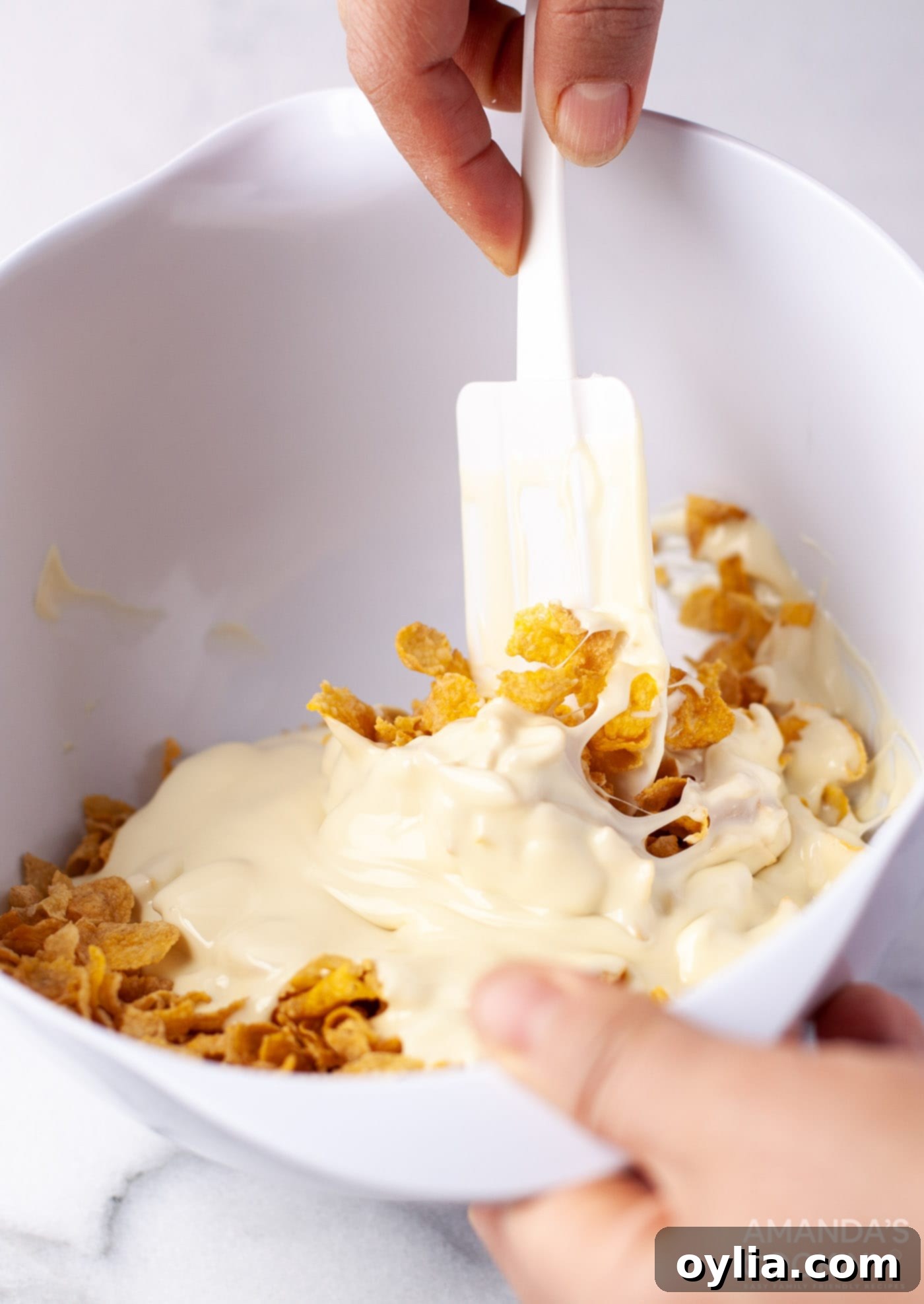 A hand gently stirring white melted chocolate into cornflakes in a bowl, ensuring even coating.