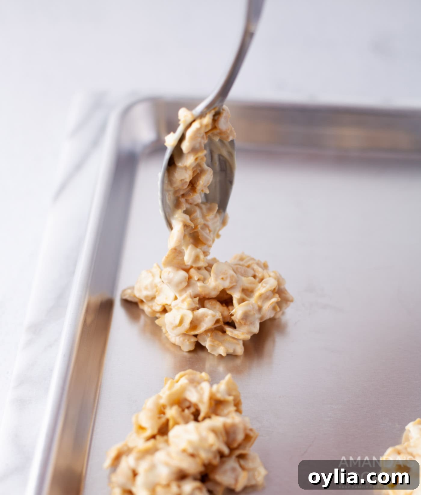 Scooping white chocolate cornflake cookie mixture onto a parchment-lined baking sheet using a tablespoon.