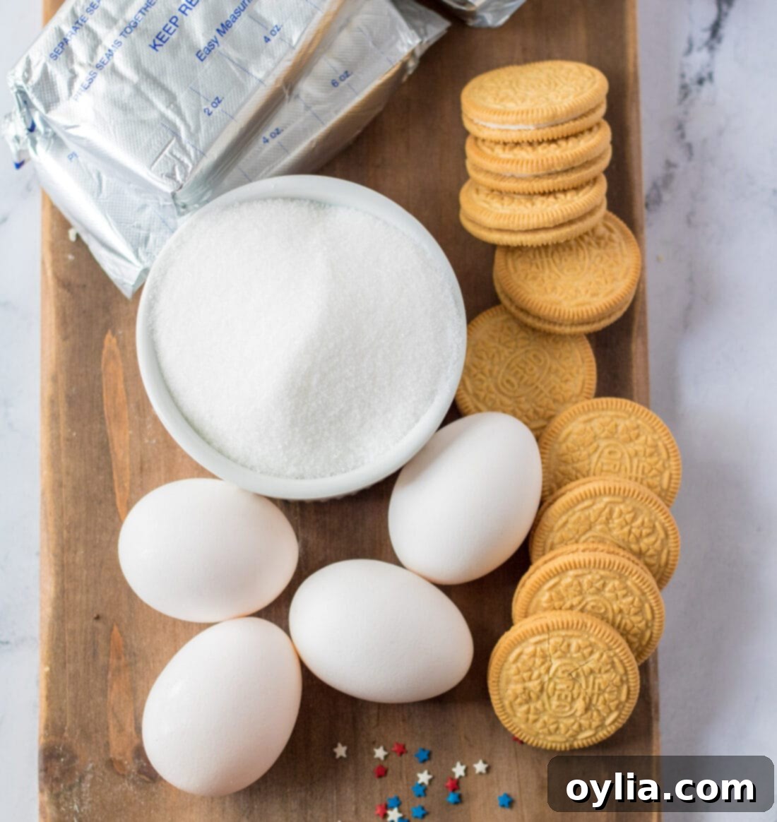 A vibrant selection of ingredients for making Patriotic Mini Cheesecakes, including blocks of cream cheese, golden Oreo cookies, a bowl of sugar, eggs, vanilla extract, and colorful sprinkles.