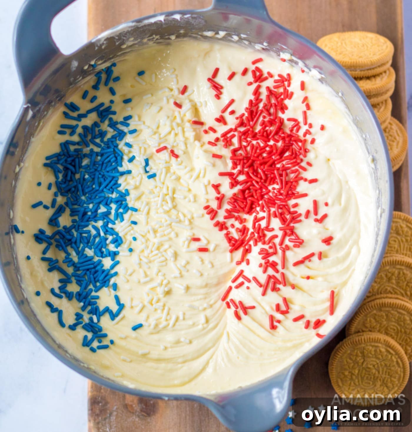 Red, white, and blue sprinkles being gently folded into creamy cheesecake batter in a bowl, ready to be poured into liners.