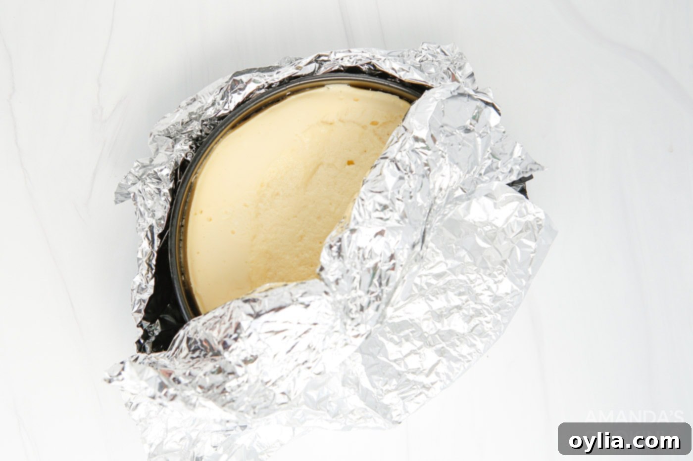 Cooked cheesecake, still in its springform pan and wrapped in aluminum foil, cooling on a counter.