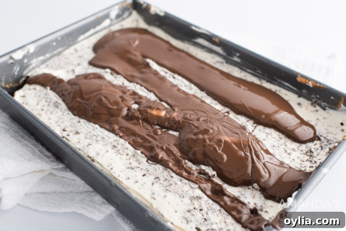 A smooth layer of chocolate ganache being poured and spread over the chilled Oreo cheesecake in a pan, with fresh Oreo crumbs ready for sprinkling.