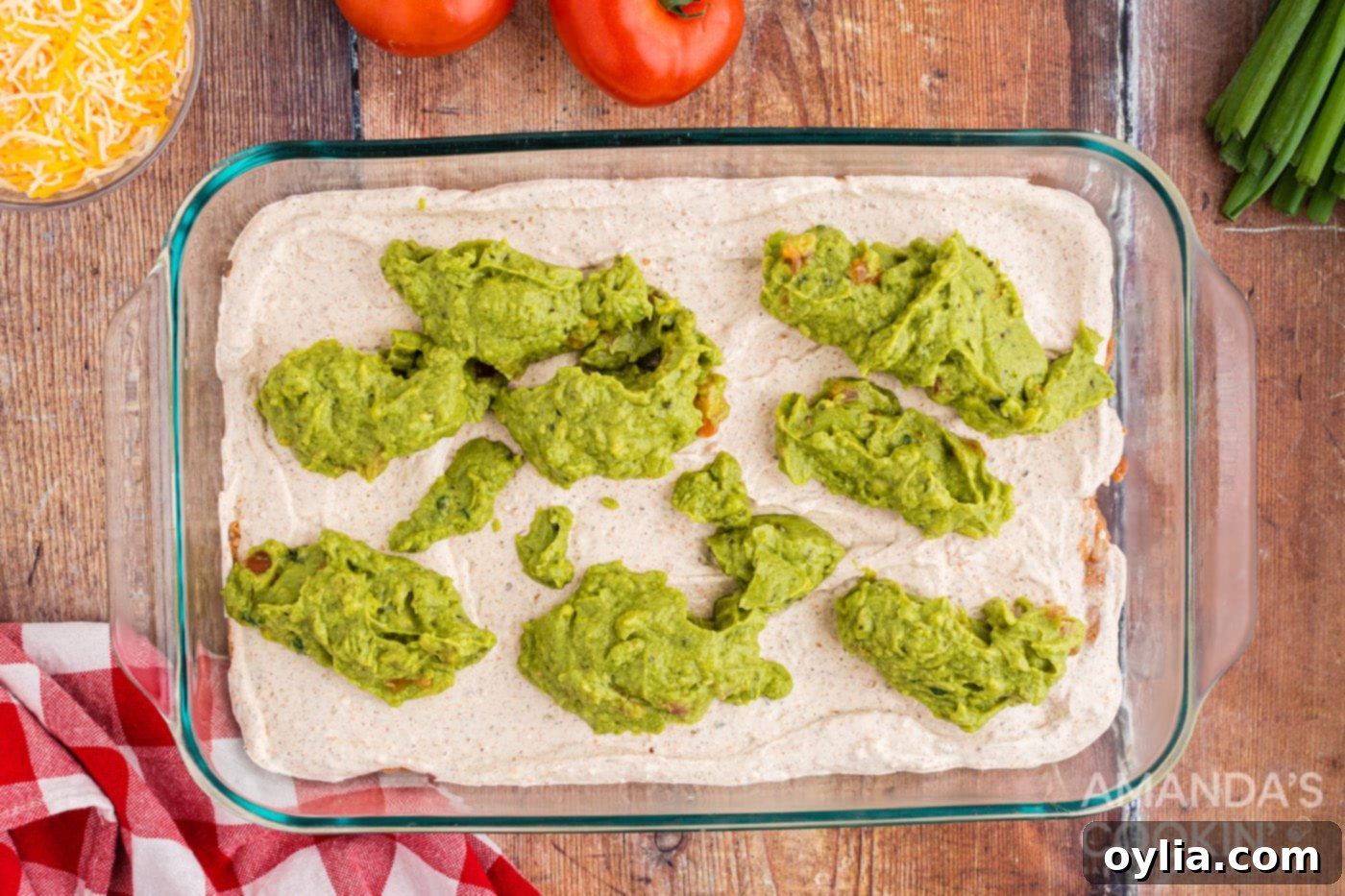 Dollops of guacamole carefully spread over the cream cheese layer in the baking dish, forming the third layer of the taco dip.