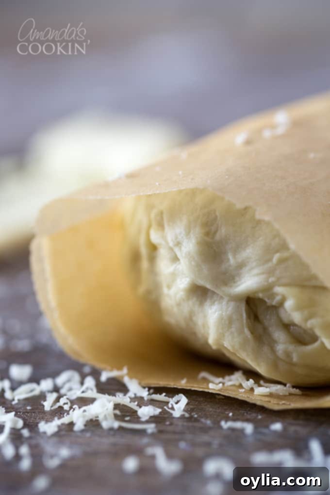 A close up photo of Parmesan garlic roll dough, generously spread with butter and cheese, then rolled up and wrapped in parchment paper, ready for chilling.