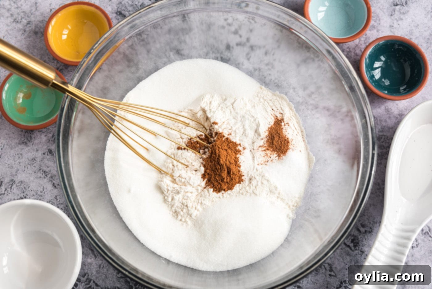 Close-up of a large bowl containing sugar, cinnamon, flour, nutmeg, and baking powder, with a whisk resting inside.