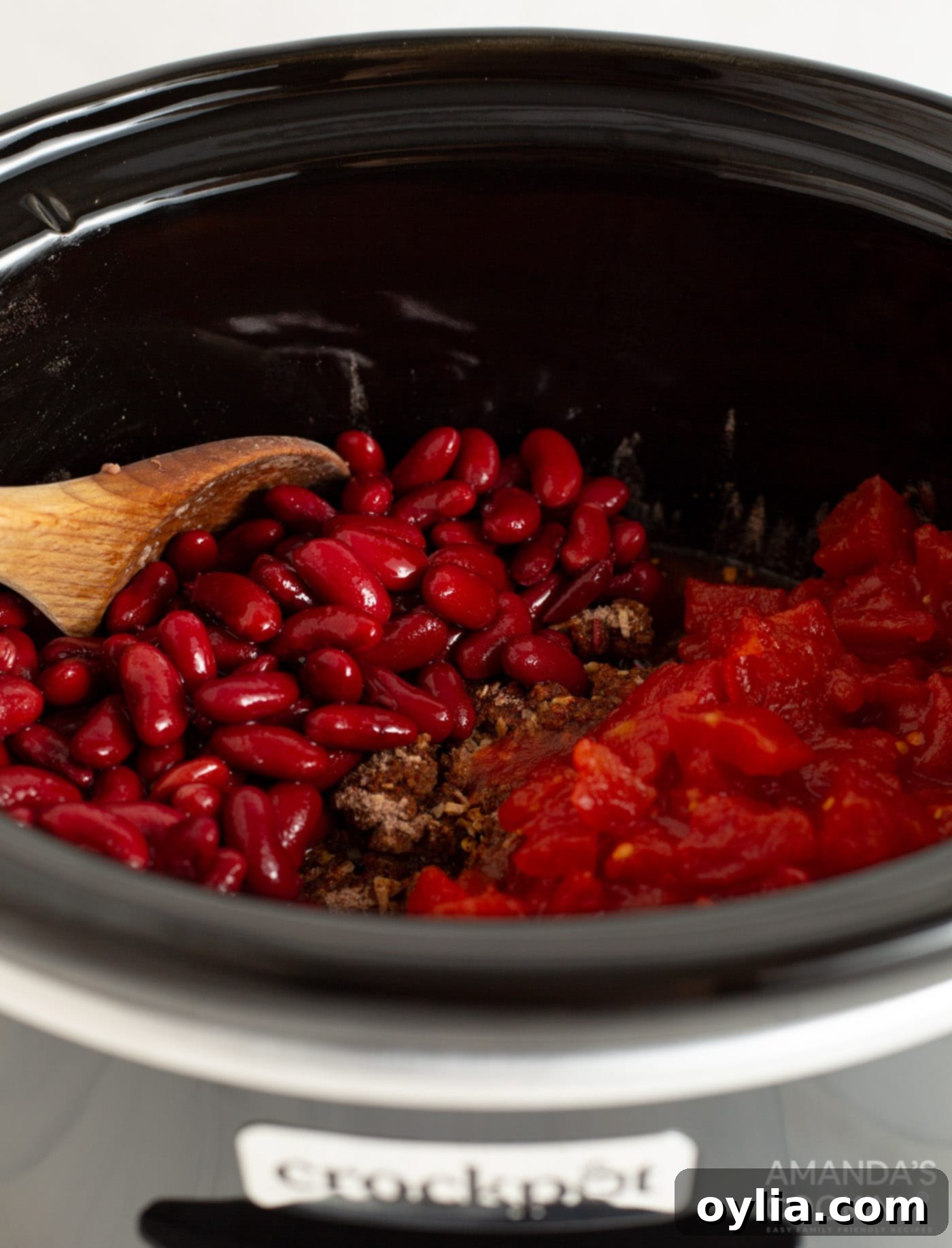 All the ingredients for a delicious Crockpot Chili, including browned beef, beans, tomatoes, and spices, fully mixed together within a slow cooker, with the lid poised to begin the slow-cooking process.