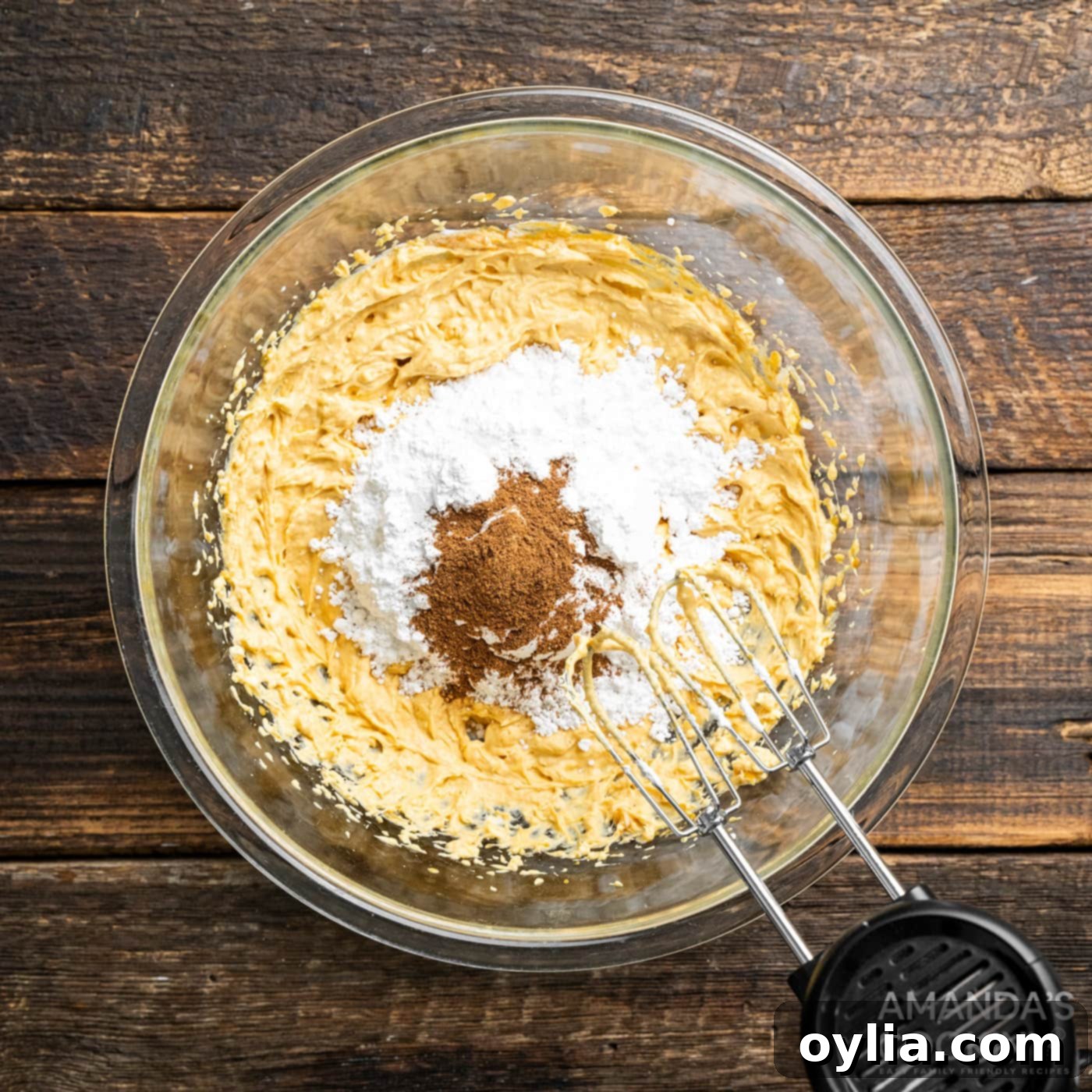 Hand mixer blending powdered sugar, pumpkin spice, and vanilla extract into the pumpkin and cream cheese mixture until smooth.