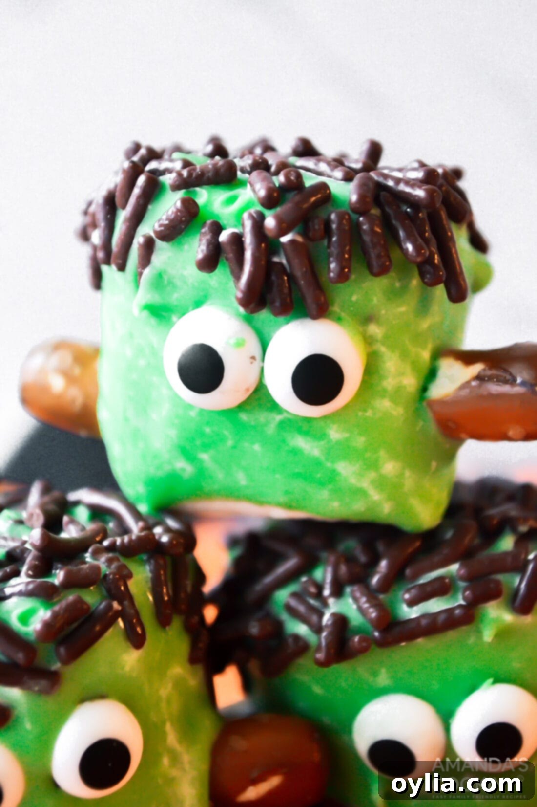 Close-up of a finished Marshmallow Frankenstein with bright green coating, black sprinkle hair, candy eyes, and pretzel bolts.
