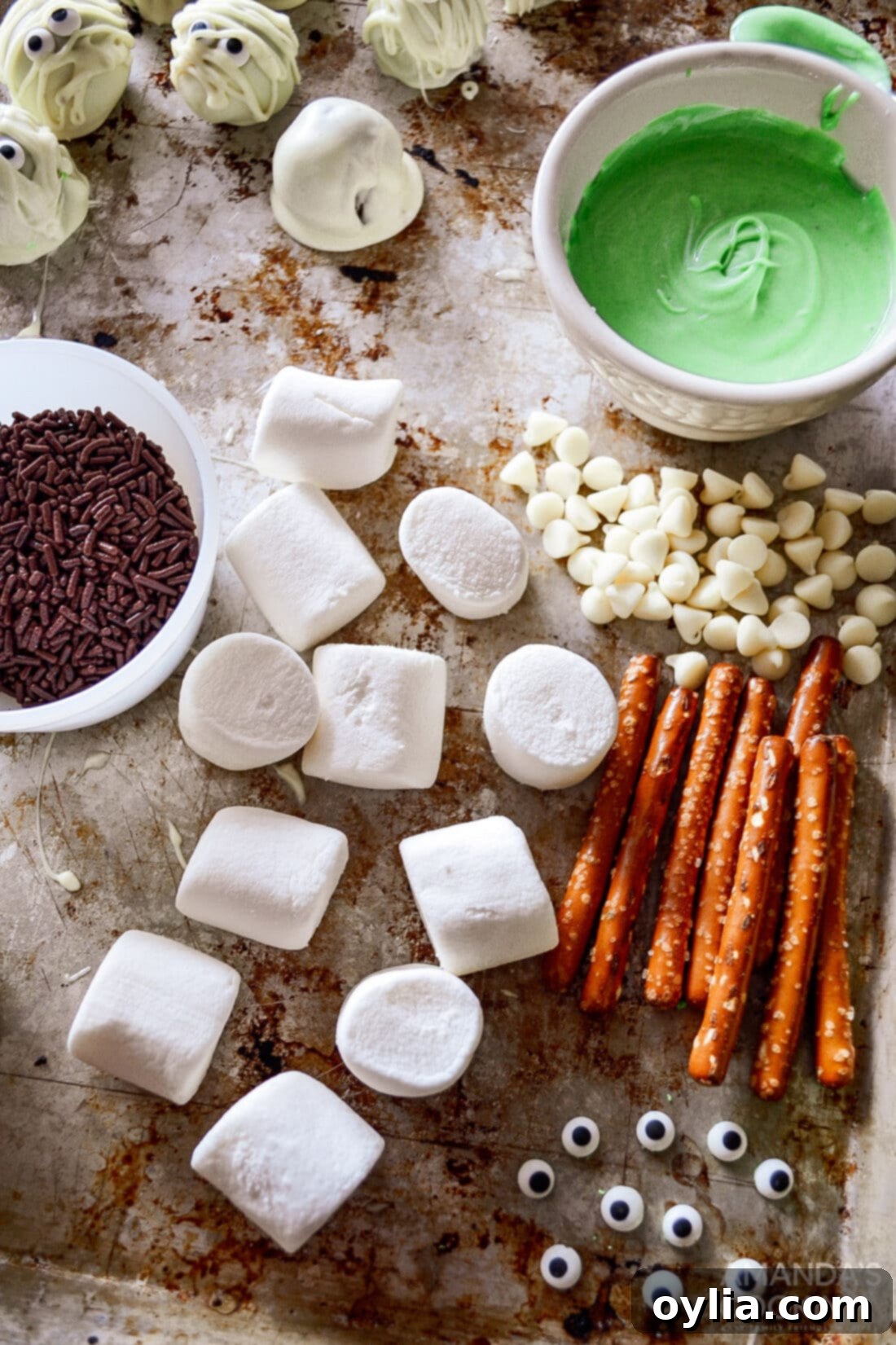 A selection of ingredients for Marshmallow Frankensteins: marshmallows, white chocolate chips, green food coloring, sprinkles, candy eyeballs, and pretzel sticks.
