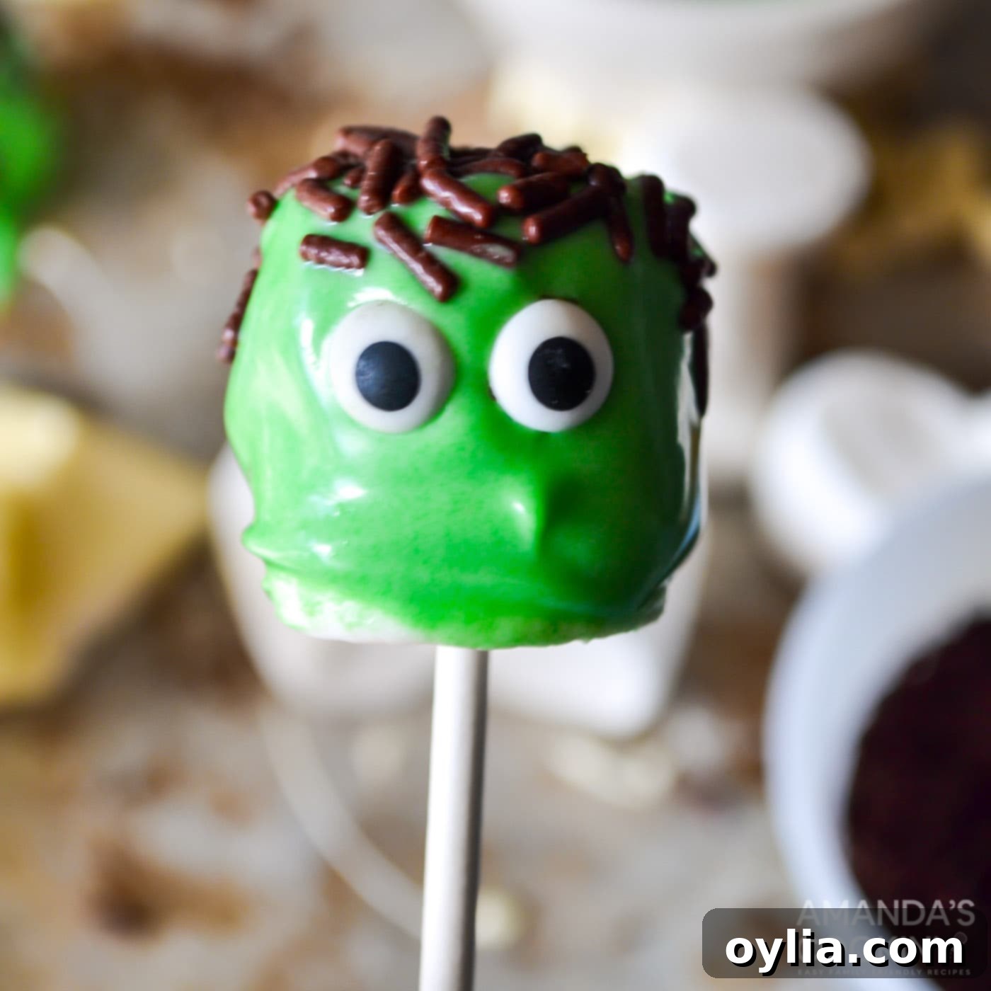 Two decorated Frankenstein marshmallow pops with green coating, sprinkle hair, and candy eyes, awaiting pretzel bolts.