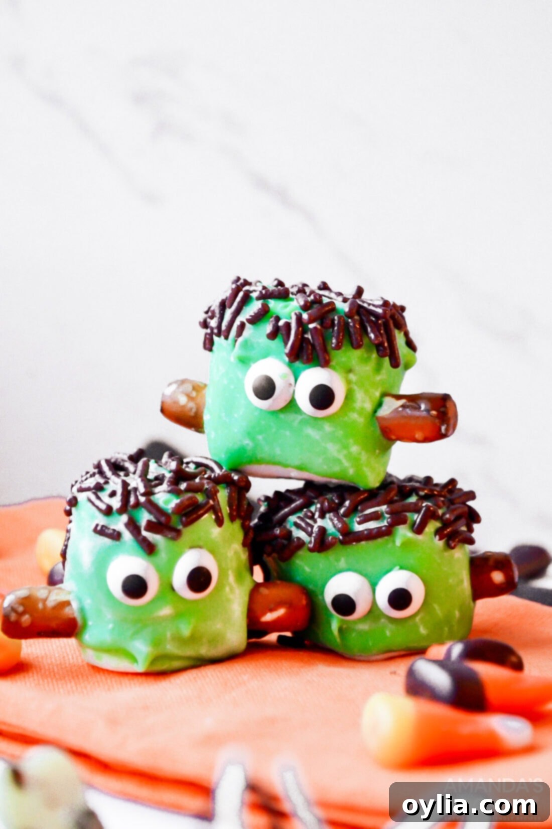 A stack of three finished Marshmallow Frankensteins, showcasing the details of the green coating, sprinkle hair, candy eyes, and pretzel bolts.