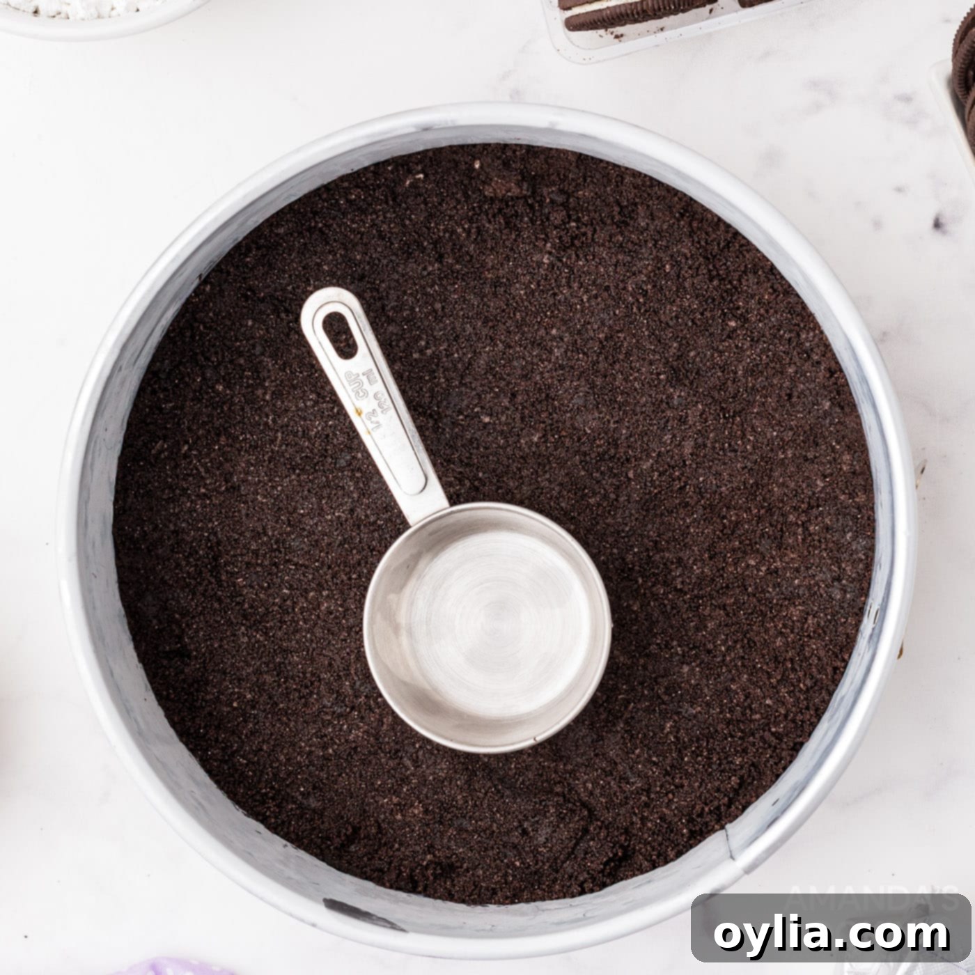 A measuring cup pressing down Oreo crumbs to form an even crust in a springform pan.