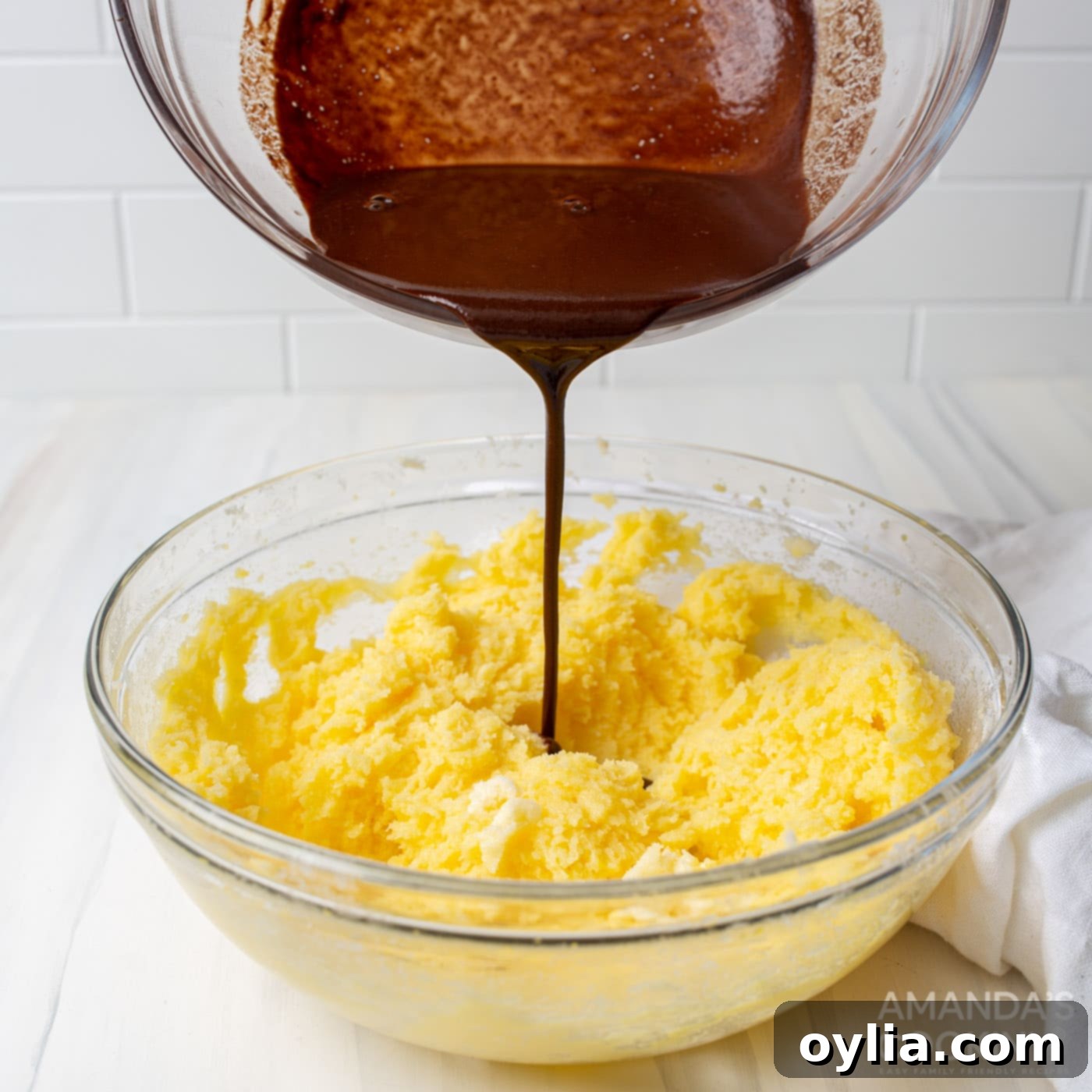 Pouring the cooled melted German chocolate into the creamy butter, sugar, and egg yolk mixture in a stand mixer.