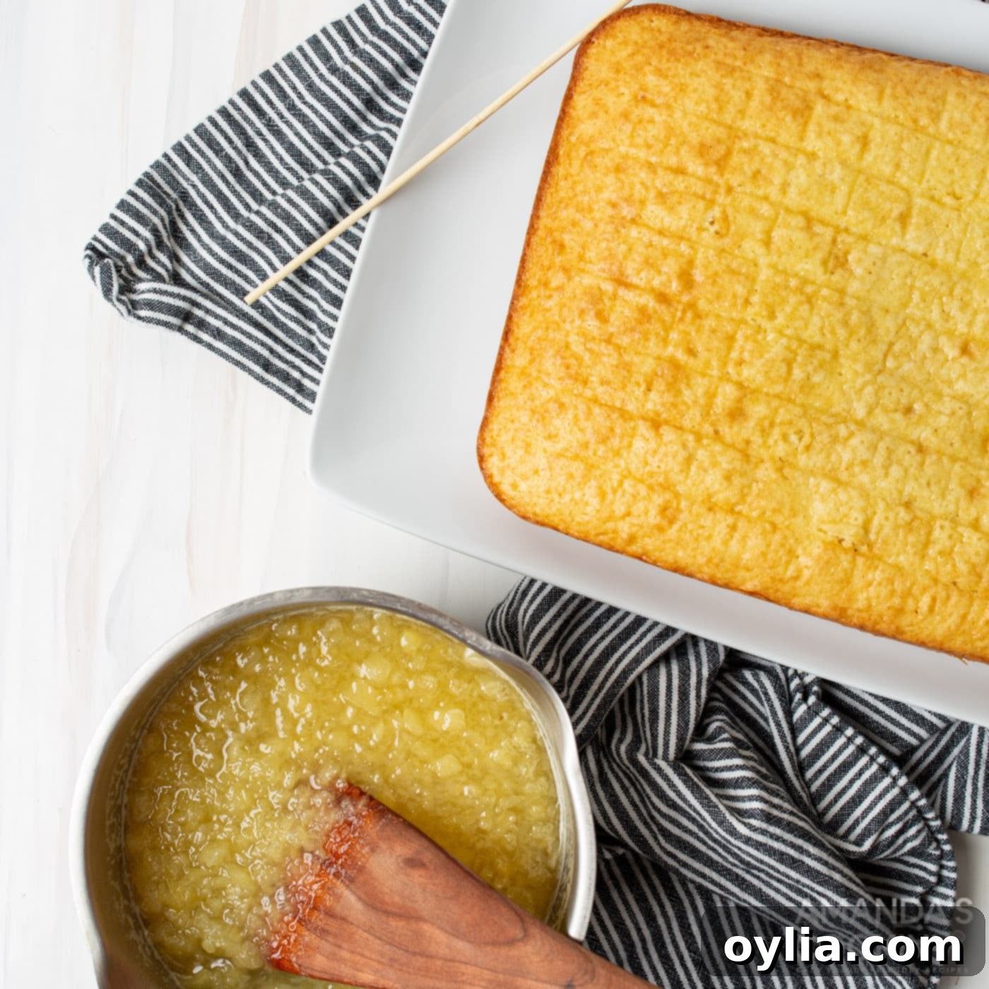 A hand pouring warm pineapple mixture over a freshly baked yellow cake with poked holes.