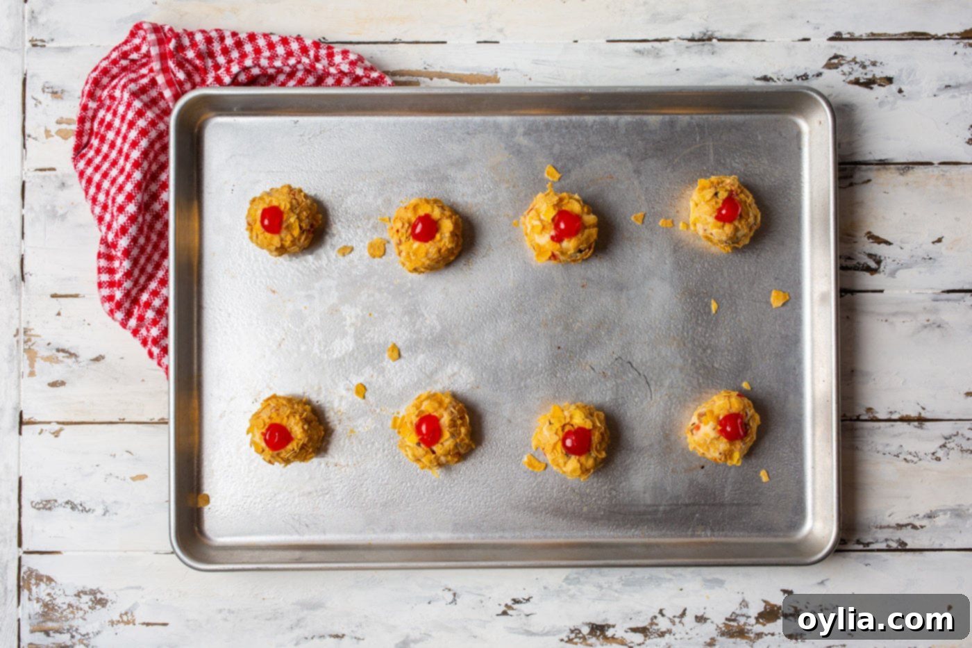 Cherry wink cookie dough balls coated in cornflakes on a baking sheet, topped with cherry pieces