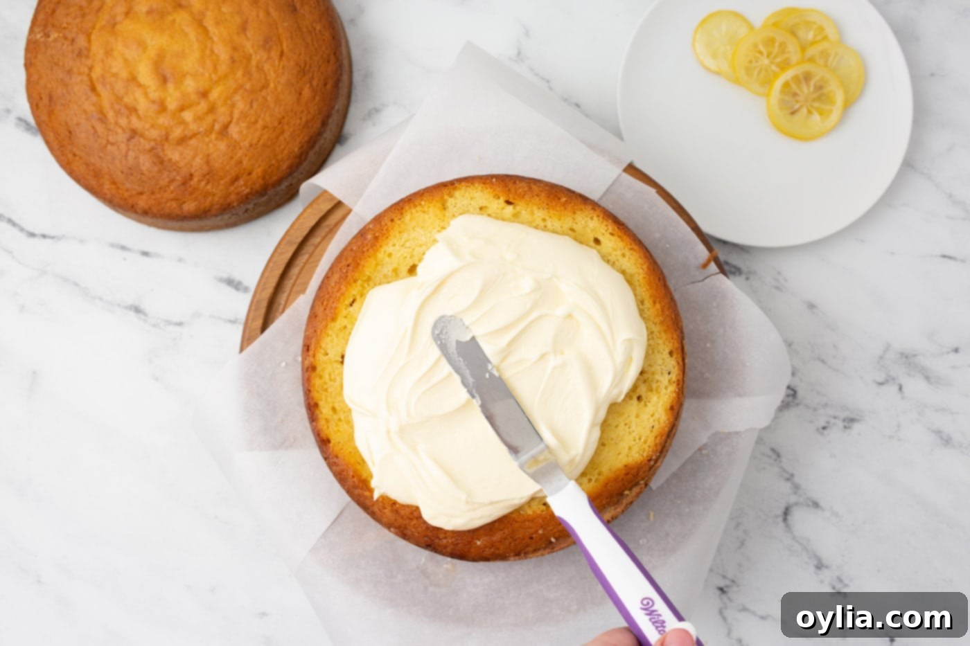 An icing spatula smoothly spreading lemon buttercream frosting onto the top layer of a lemon cake, demonstrating the frosting technique.