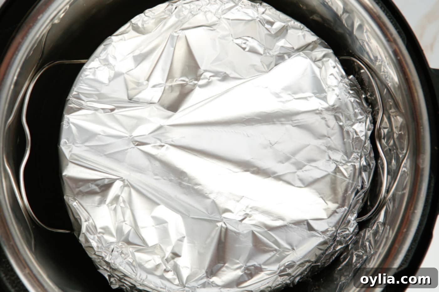 Aluminum foil-wrapped cheesecake being carefully lowered into the Instant Pot on a trivet