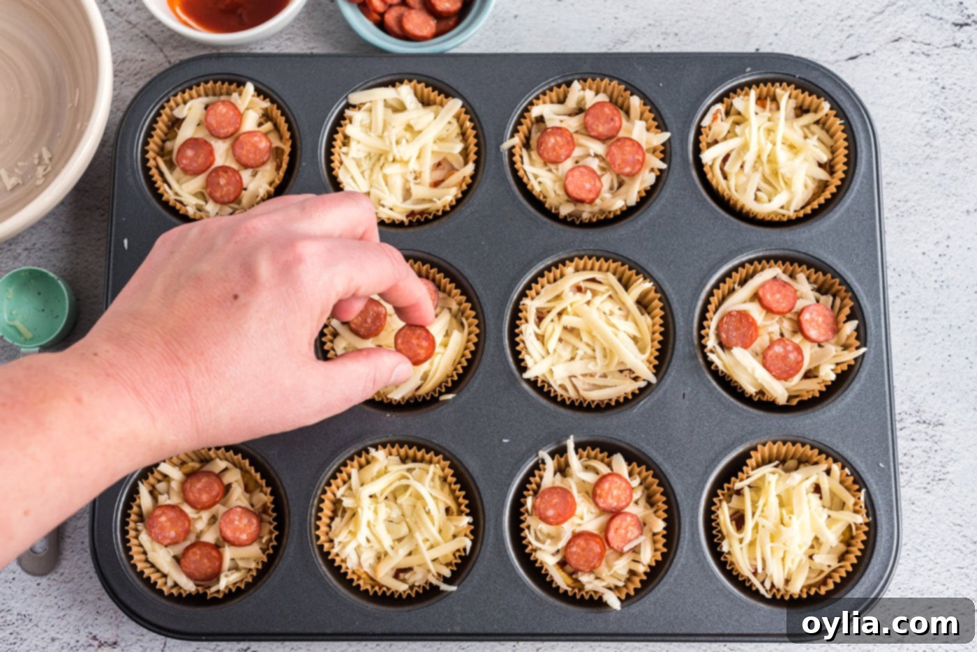 A hand placing mini pepperoni slices onto a cauliflower pizza bite topped with cheese and sauce.