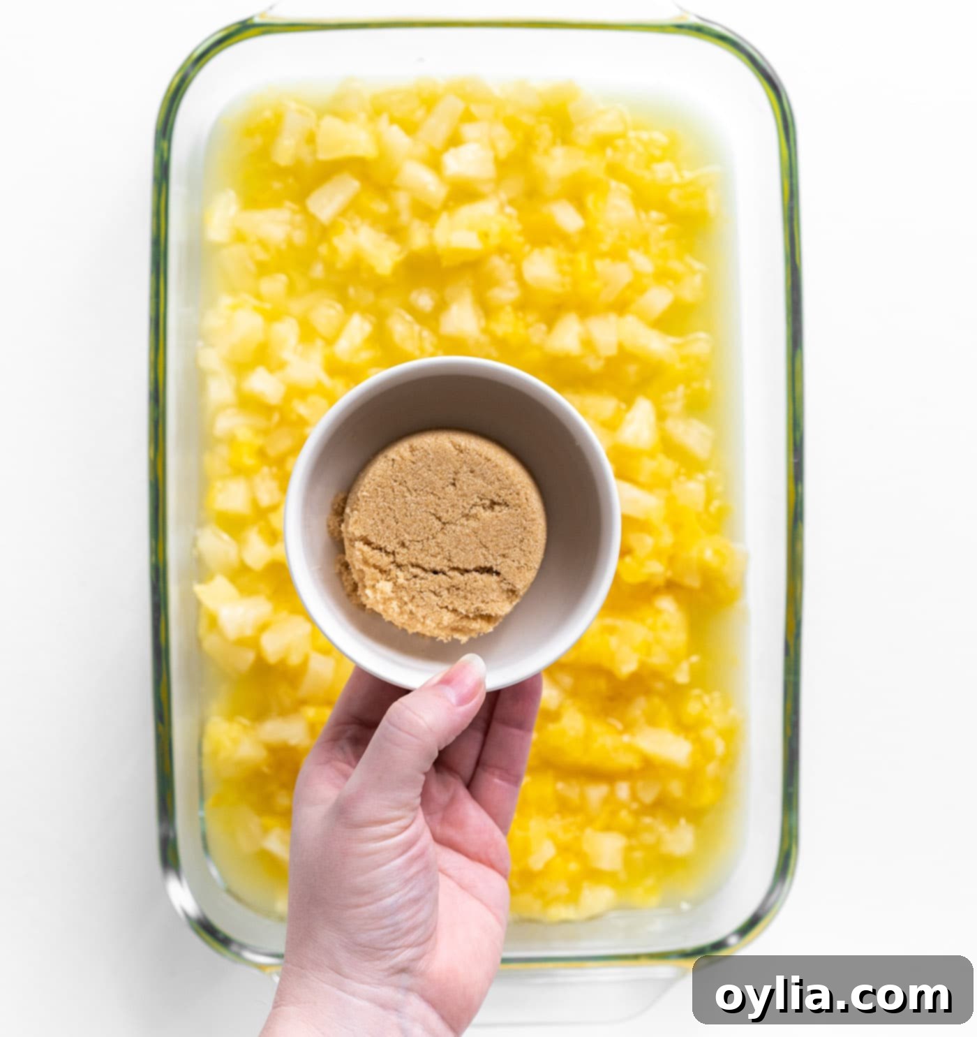 Hand sprinkling light brown sugar over crushed pineapple in a baking dish