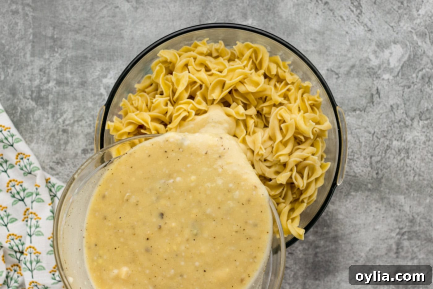 pouring sauce over egg noodles, tuna, cheese, and vegetables in a bowl
