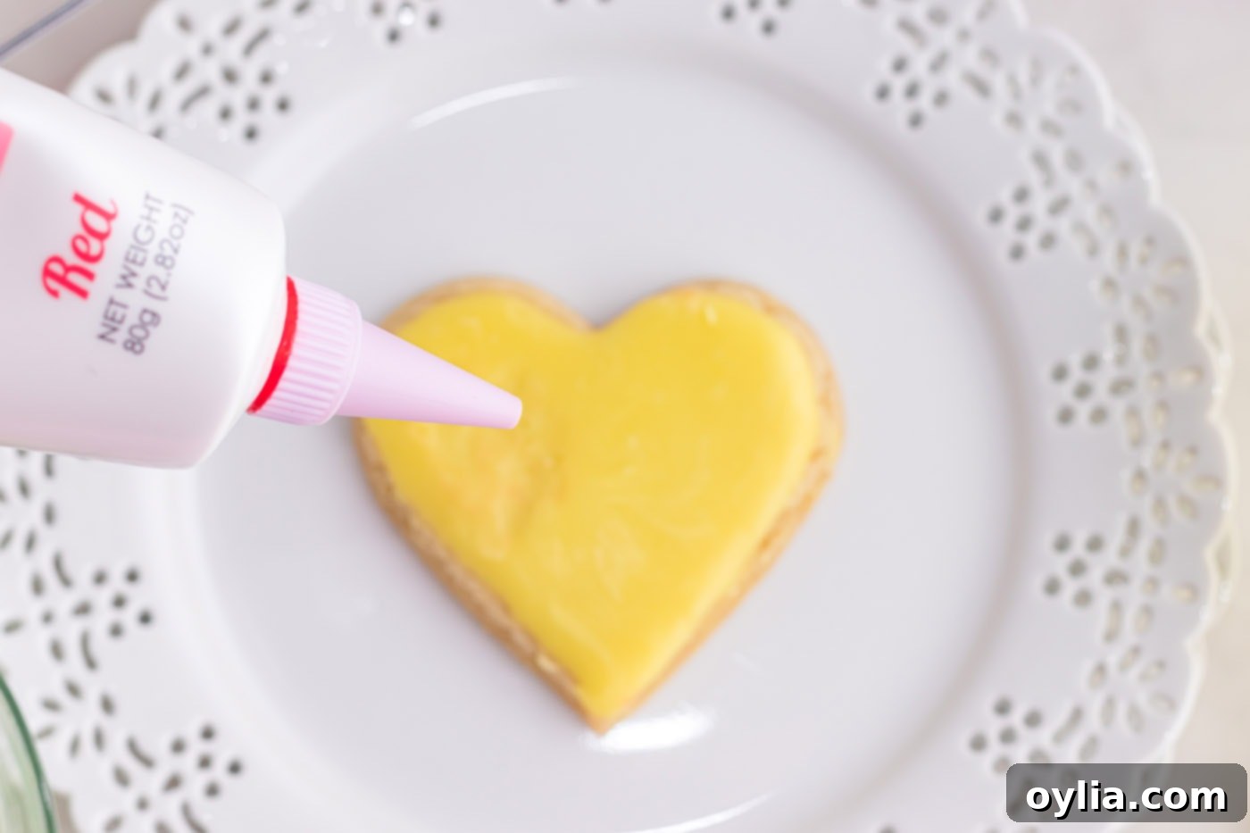 A hand carefully piping a red gel frosting message onto a pink heart-shaped cookie.