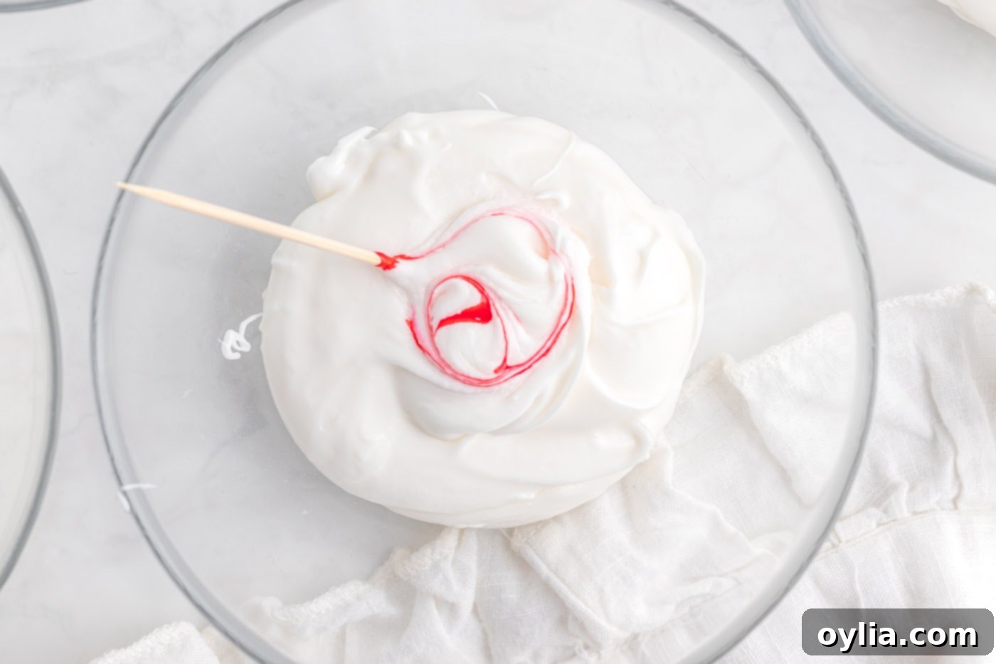 A toothpick gently mixing red gel food coloring into a bowl of white frosting to create a soft pink hue.