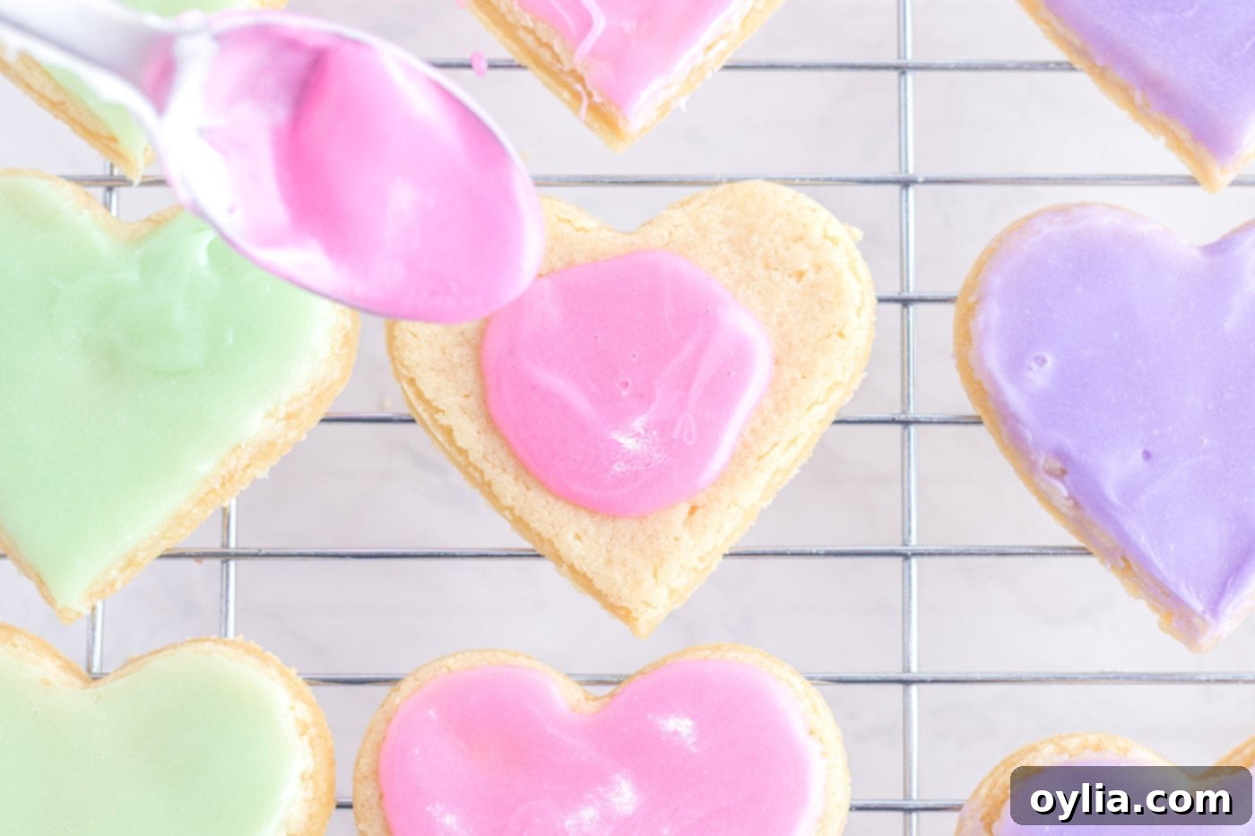 A spoon dolloping smooth pink frosting onto a heart-shaped sugar cookie, ready to be spread evenly.